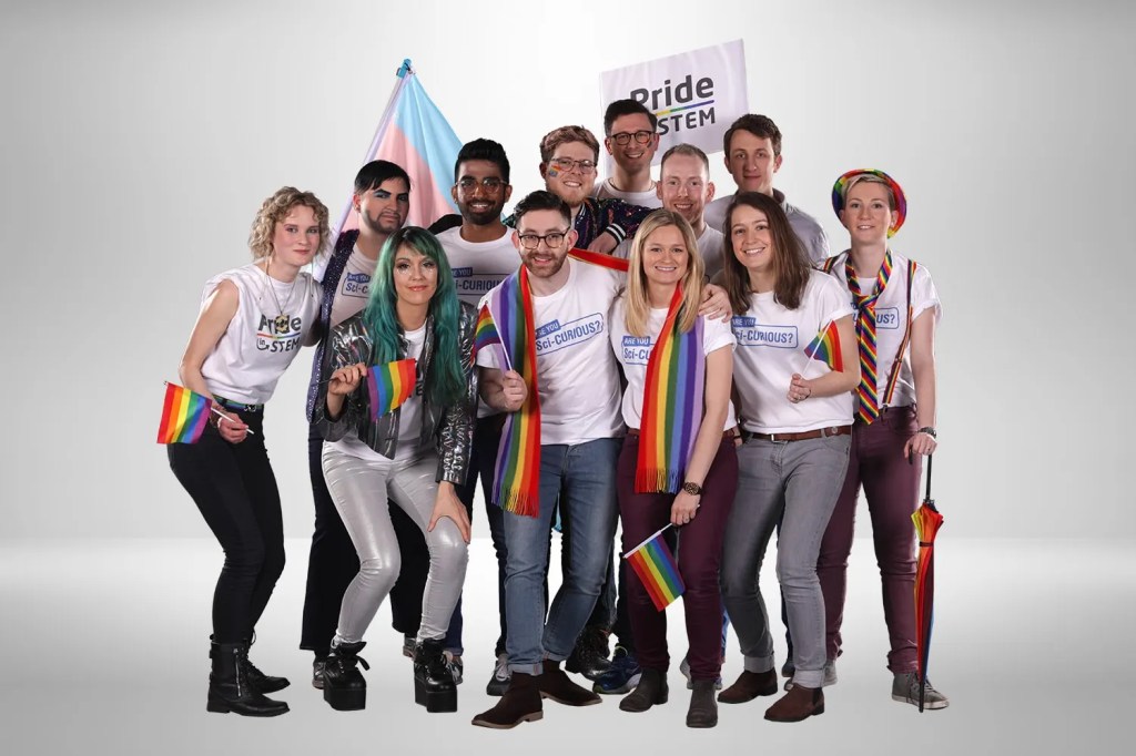 A photo of several people against a grey-white background. The people have their arms around each others shoulders, most are wearing white shirts that say "Pride in STEM" and "Sci-curious" and they are all waving rainbow-coloured pride flags. An actual trans flag is flying in the background, and one person is holding a placard that says "Pride in STEM".